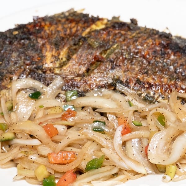 Whole grilled fish with crispy skin, served with sautéed onions, peppers, and a side of red sauce.